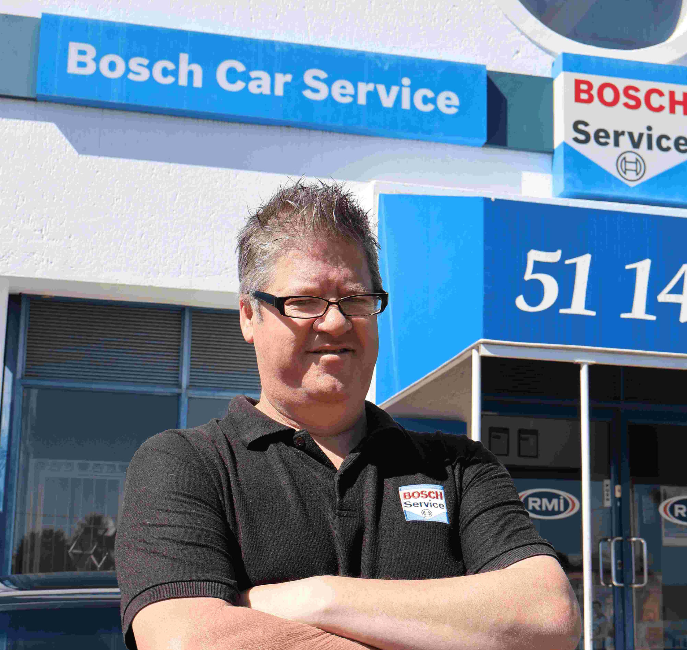 Manager Jacques Petzer with over 20 years of experience, standing with arms folded, wearing Bosch Service shirt, standing outside of Bosch Car Service Northcliff Auto. RMI logo in the background, on the glass doors of the building.